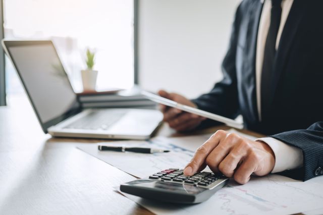 Image d’un homme d’affaires assis à un bureau avec des documents, un ordinateur portable ouvert et tapant sur une calculatrice.